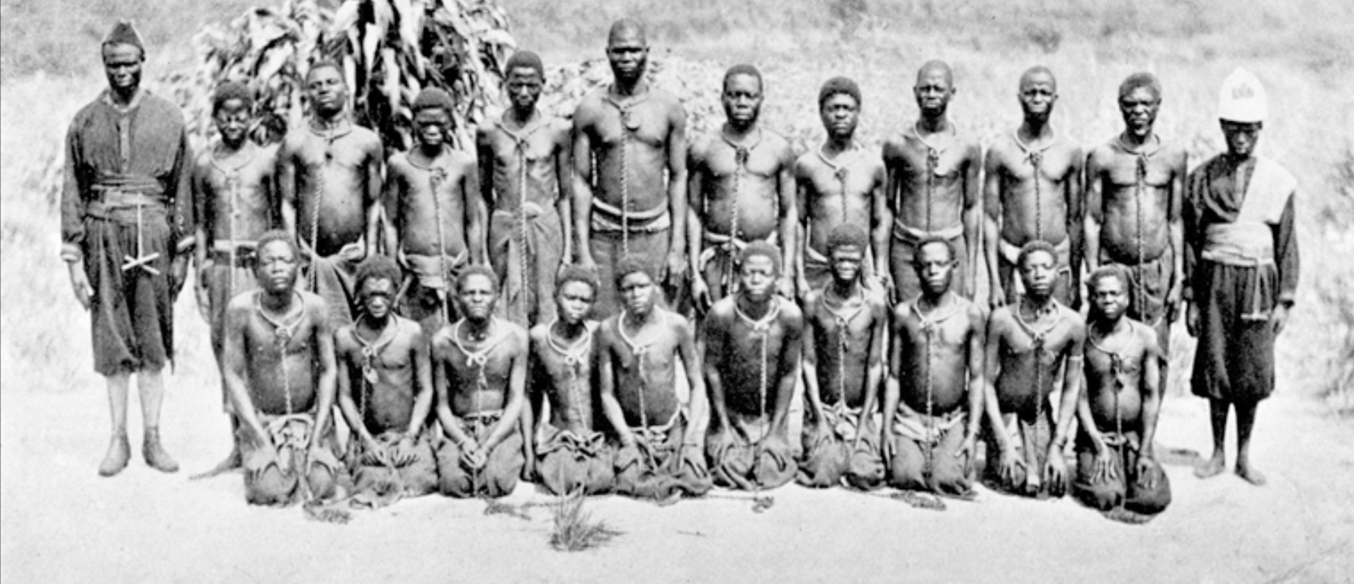 Two rows of prisoners with chains around their necks, flanked by two guards.