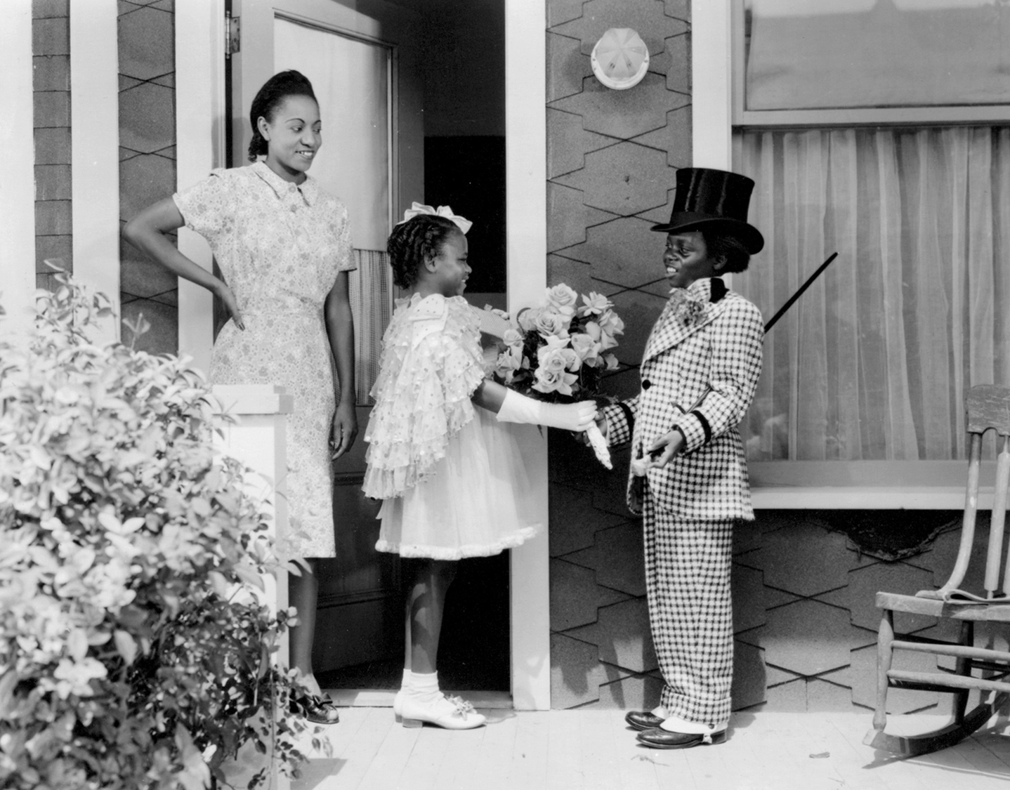 Thomas as the Buckwheat character dressed in a suit with cane shaking a young girl's hand on the porch while a woman smiles in the background