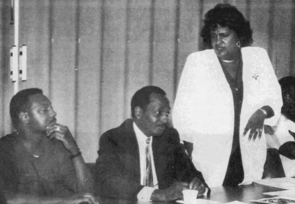 Bajoie standing and speaking while Jones and Jackson sit at a desk