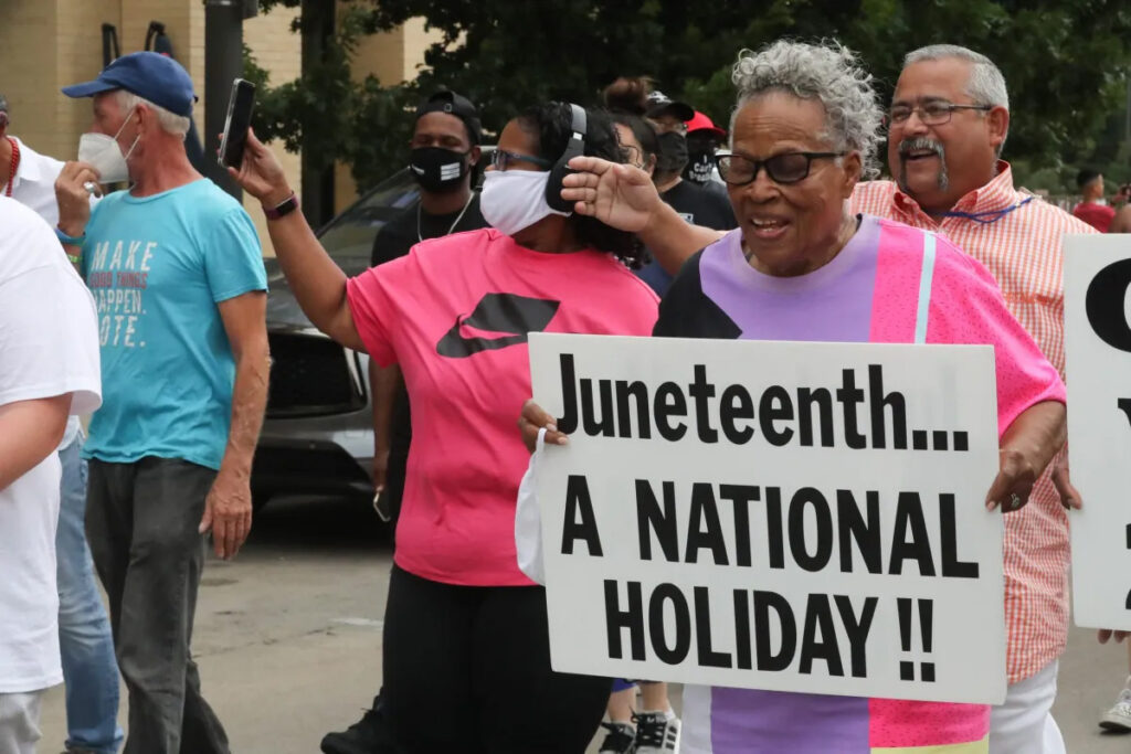 Lee with a group of others on the street, holding a sign reading "Juneteenth... A National Holiday"