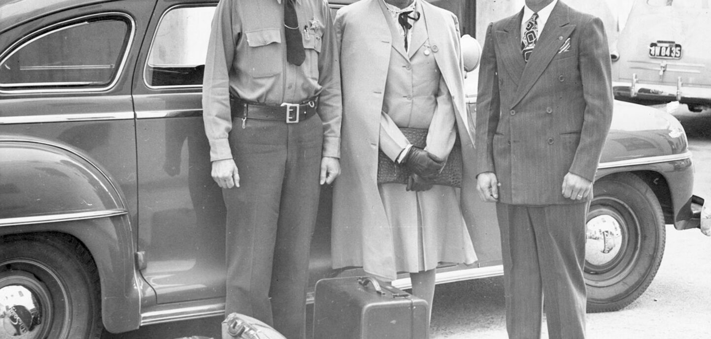 Lucy Hicks stands with two deputy sheriffs in front of an automobile. Left to right: Deputy Sheriff (in uniform with hat) H.E. Bowman, Lucy Hicks Anderson, Deputy Sheriff Charles Salig. Lucy Hicks wears a skirt suit with a coat draped over her shoulders and a hat, and Charles Salig wears a suit, tie, and hat. Three pieces of luggage lay at their feet.