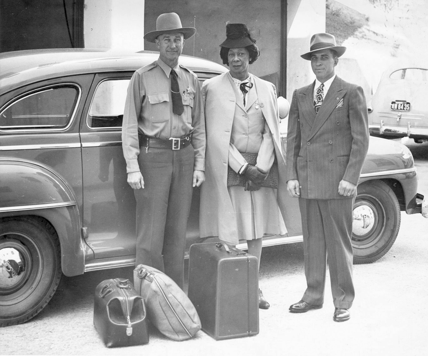 Lucy Hicks stands with two deputy sheriffs in front of an automobile. Left to right: Deputy Sheriff (in uniform with hat) H.E. Bowman, Lucy Hicks Anderson, Deputy Sheriff Charles Salig. Lucy Hicks wears a skirt suit with a coat draped over her shoulders and a hat, and Charles Salig wears a suit, tie, and hat. Three pieces of luggage lay at their feet.