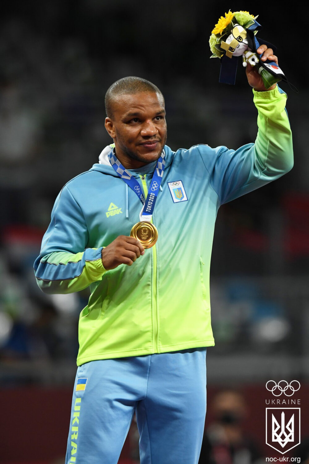 Beleniuk in a green and blue tracksuit holding a bouquet of flowers and presenting a gold medal on a lanyard