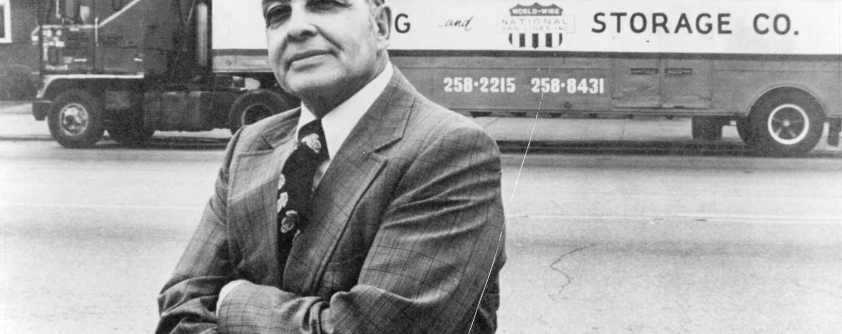Ward with arms crossed in a suit and tie in front of a truck that reads "E.E. Ward Storage Co."