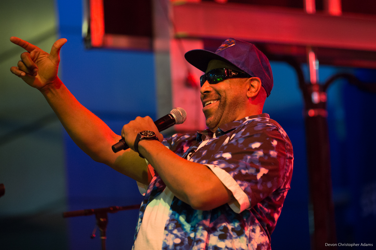 Tone Loc smiling in sunglasses and baseball cap holding a microphone and pointing to the audience