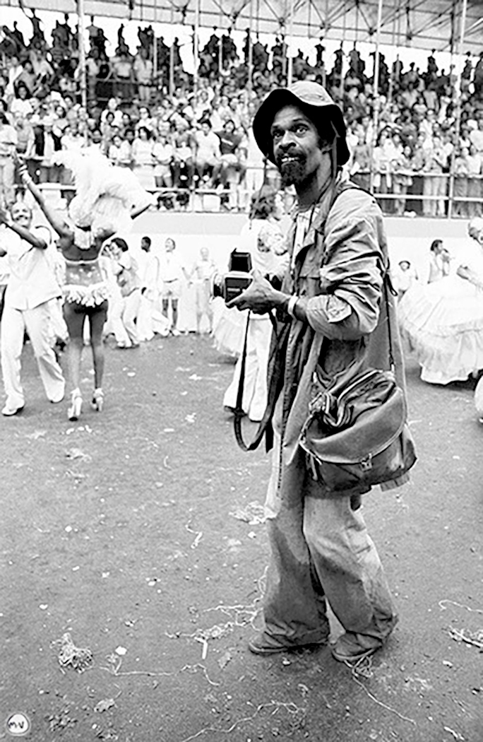 Garcia in a bucket hat, loose fitting clothes and a leather side bag holding a camera towards costumed Carnival dancers being watched by an audience in stadium seating in the background