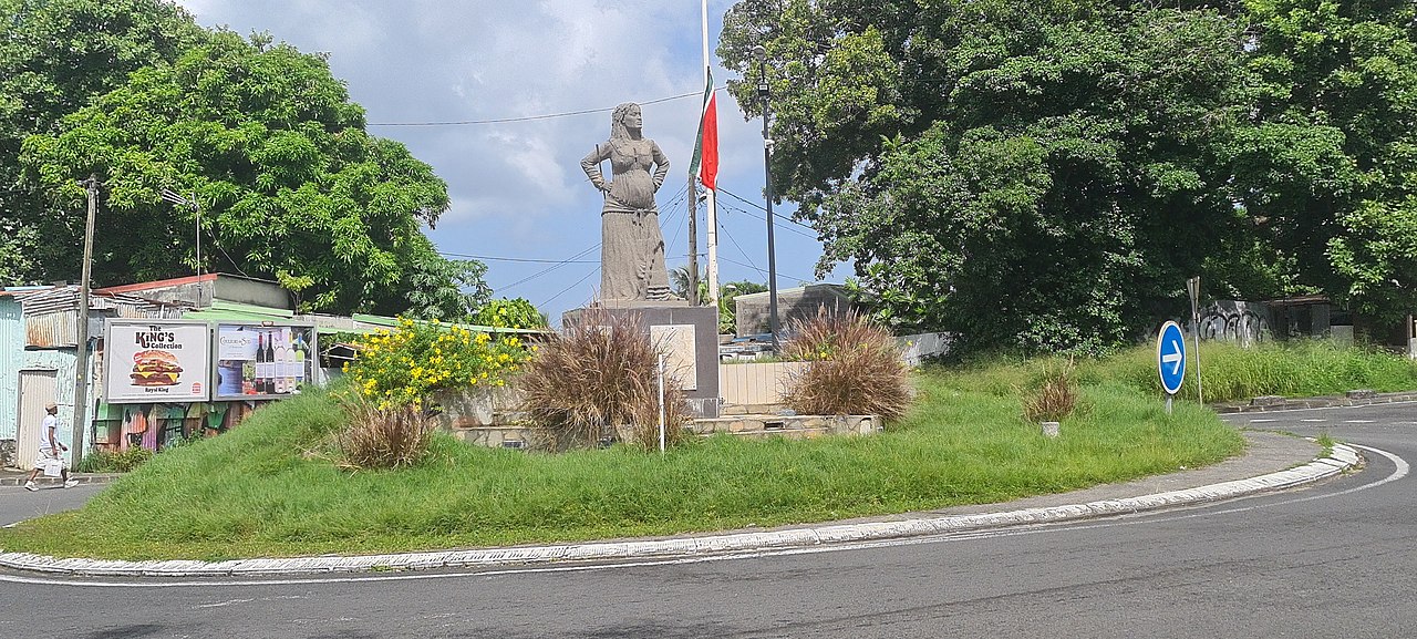 Statue in the middle of a roundabout next to flowers with flag at half mast and billboards in the background