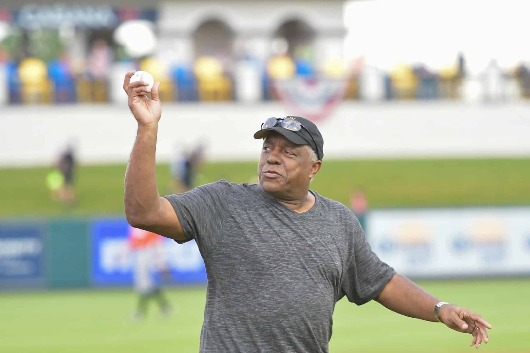 An older LeFlore holding a baseball in the pitching position, wearing a black t-shirt baseball cap and sunglasses over the brim