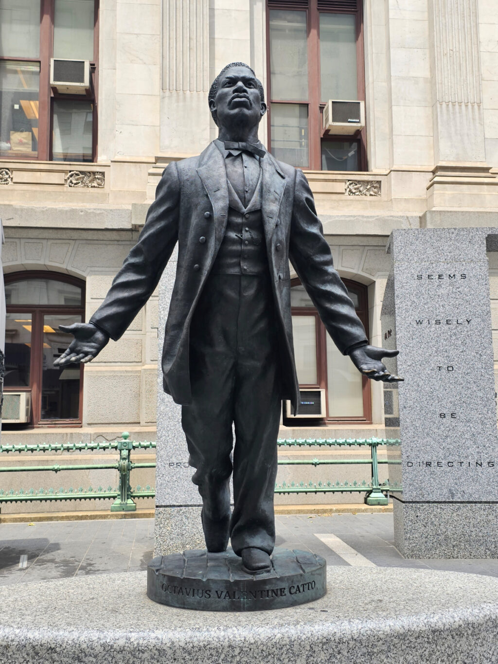 Statue of Catto in formal wear and bowtie with arms outstretched beside a rectangular column reading "seems wisely to be directing"