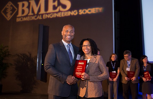 Ameer smiling beside plaque and curtain reading "BMES Biomedical Engineering Society"